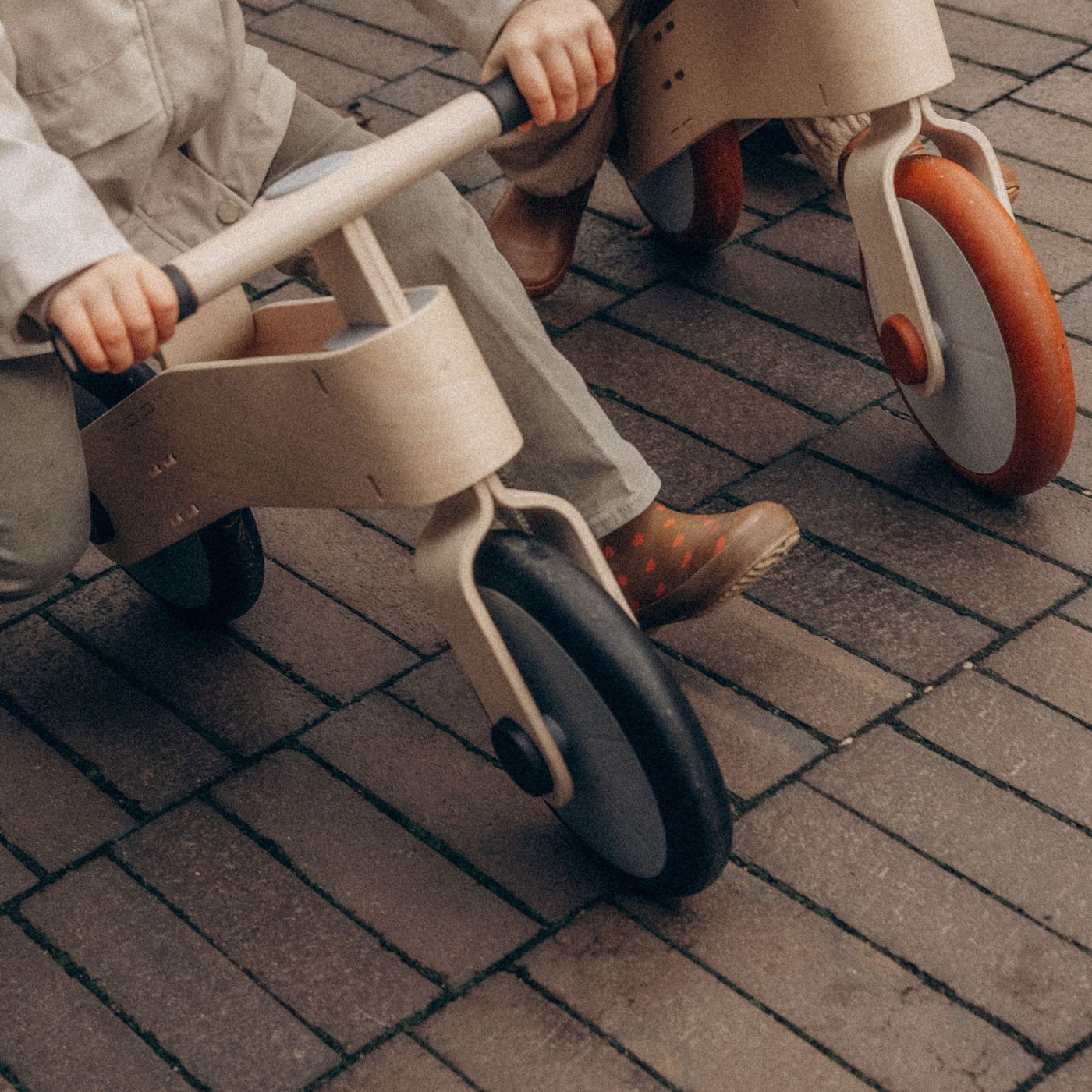 Toddler riding a wooden balance bike next to another child on a brick path